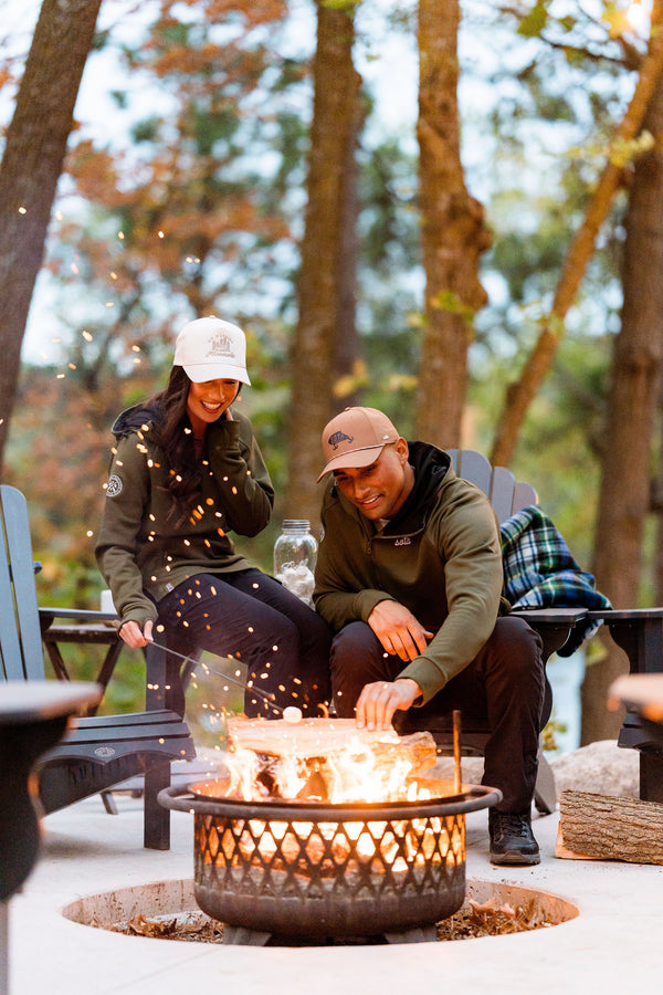 Two people sitting around a fire pit in a forest setting wearing sota clothing 