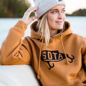 Person wearing a brown hoodie with 'SOTA' logo and a gray beanie by a lake.