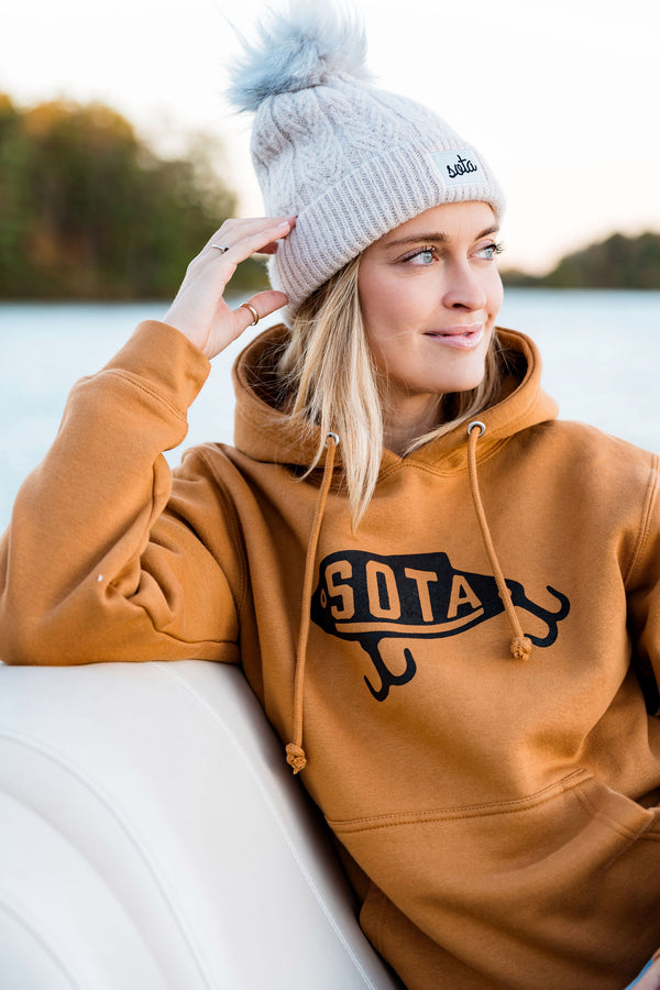 Person wearing a brown hoodie with 'SOTA' logo and a gray beanie by a lake.