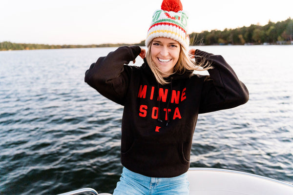 Person wearing a 'MINNESOTA' sweatshirt on a boat with a Minnesota Wild Hockey Team inspired beanie. There is a lake in the background.