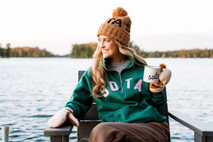 Person sitting by a lake wearing a green sweatshirt with 'Sota' text, brown beanie, and holding a mug.