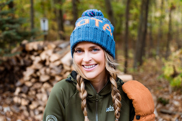 Woman wearing a blue knit hat with 'sota beanie' text, green jacket, and carrying a brown backpack in a forest setting.