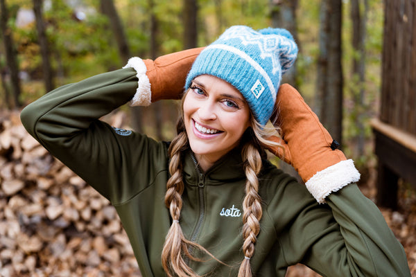 Woman wearing a blue beanie and green jacket with 'sota' logo in a forest setting