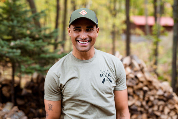 Man wearing a green cap with a Minnesota logo in a forest setting
