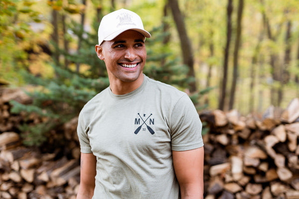 Man wearing a green t-shirt with a logo and a white cap, standing in a forest.
