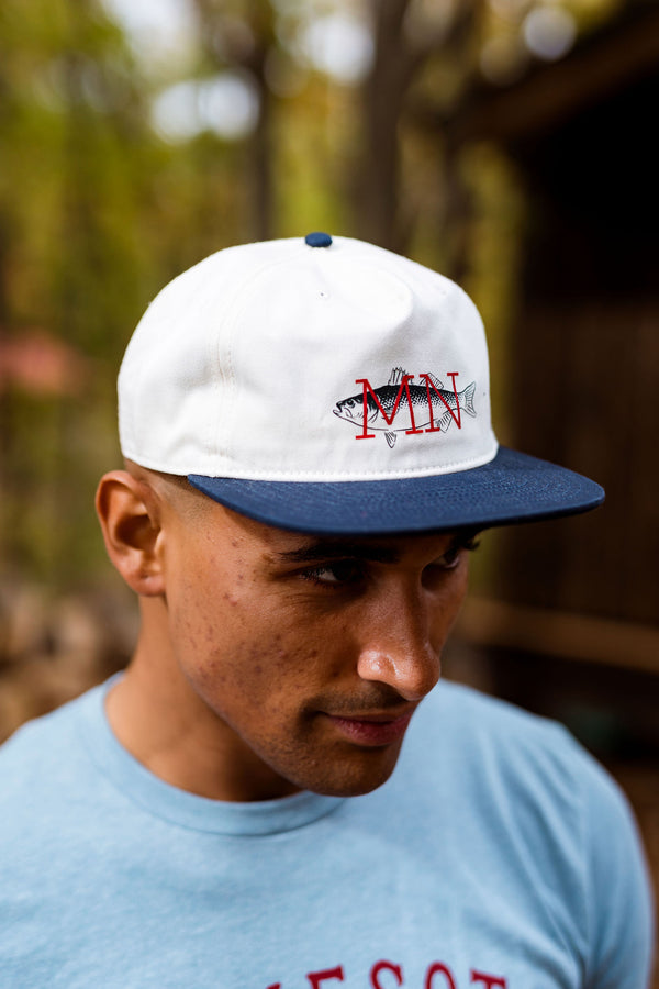 Person wearing a white and blue cap with a Minnesota fish logo, outdoors.