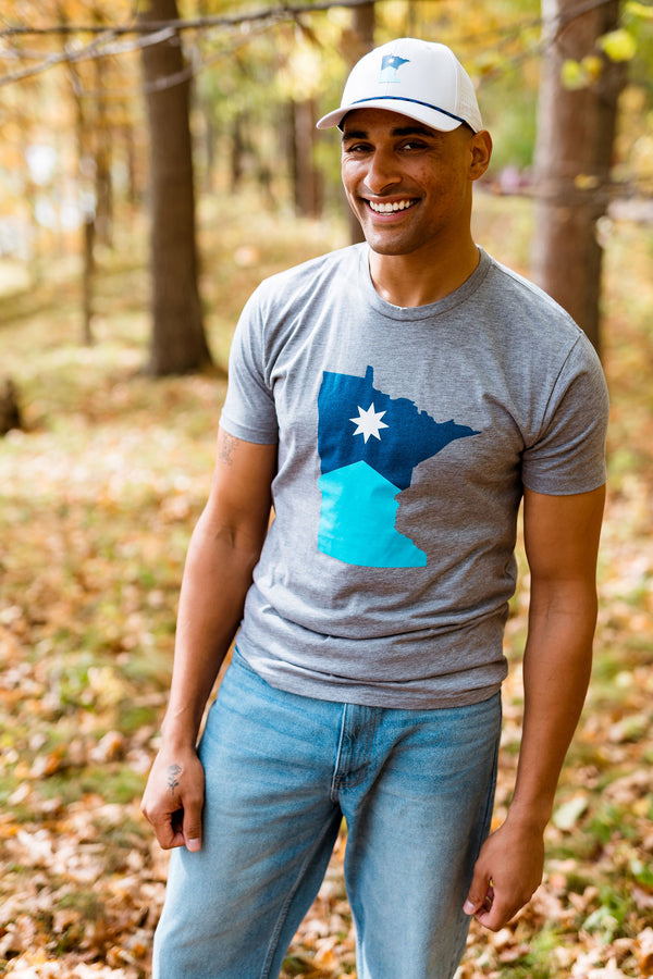 Man wearing a gray t-shirt with a blue graphic of the state of Minnesota, standing in a forest.