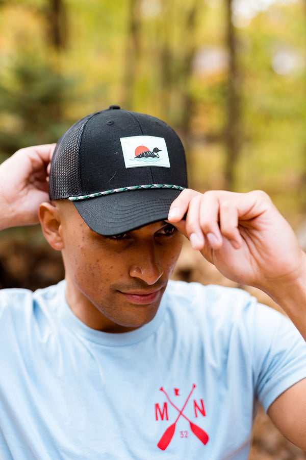Person wearing a black cap with a loon logo in a forest setting