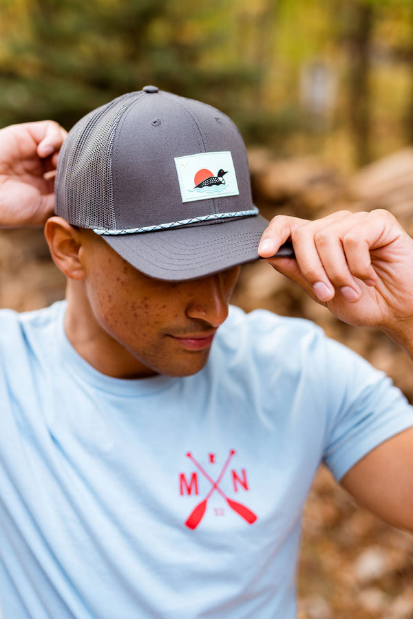 Person wearing a gray cap with a loon logo and a light blue t-shirt with a red minnesota logo in a forest setting