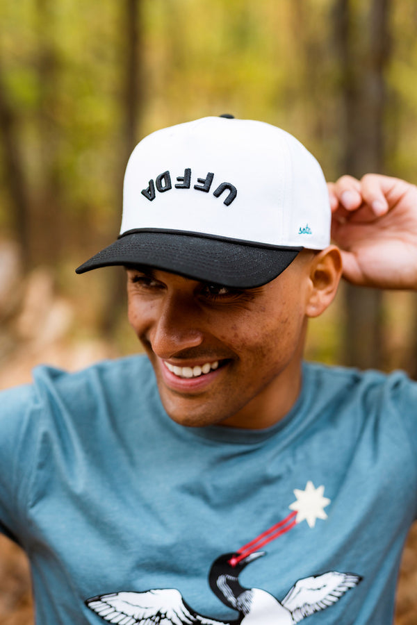 Man wearing a cap with an upside down UFFDA logo in a forest setting