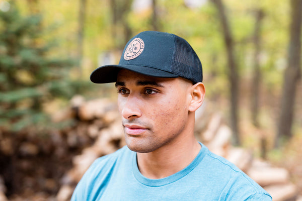Man wearing a blue t-shirt with a laser loon graphic design and a minnesota-centric black cap with hockey sticks  in a forest setting