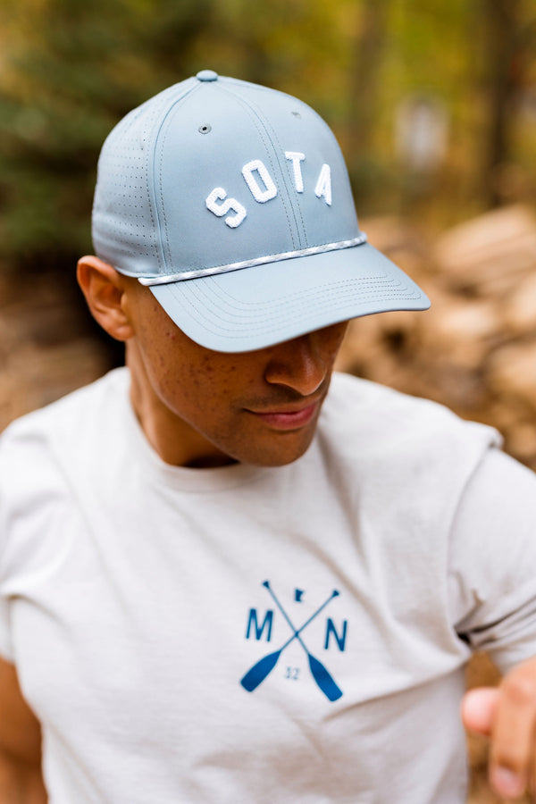 Person wearing a light blue cap with 'SOTA' and a white t-shirt with a logo, outdoors.