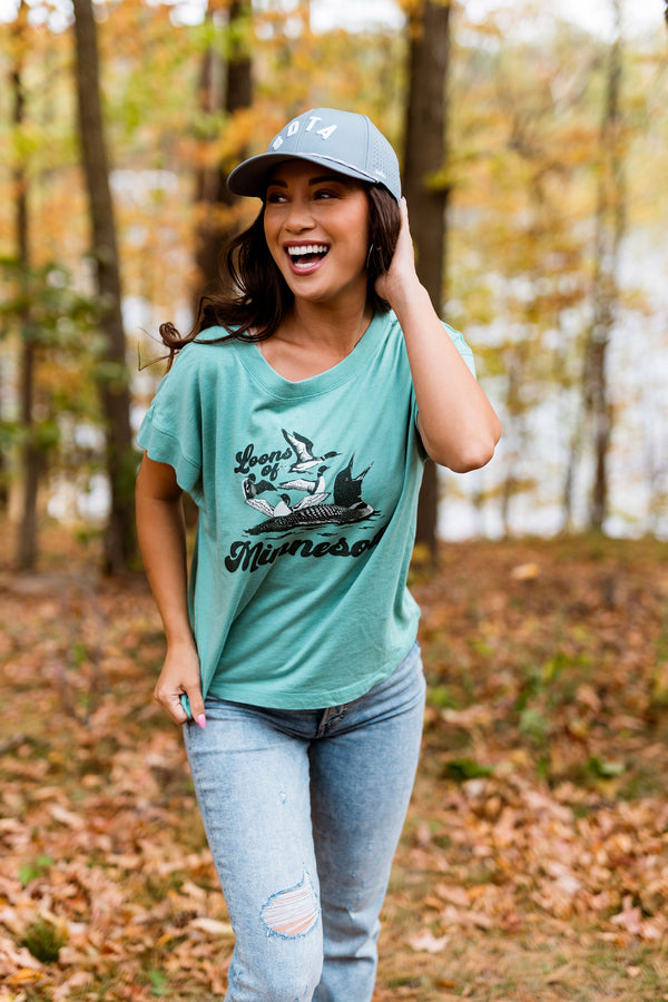 Woman wearing a green t-shirt with a graphic design and a cap, standing in a forest with autumn foliage.