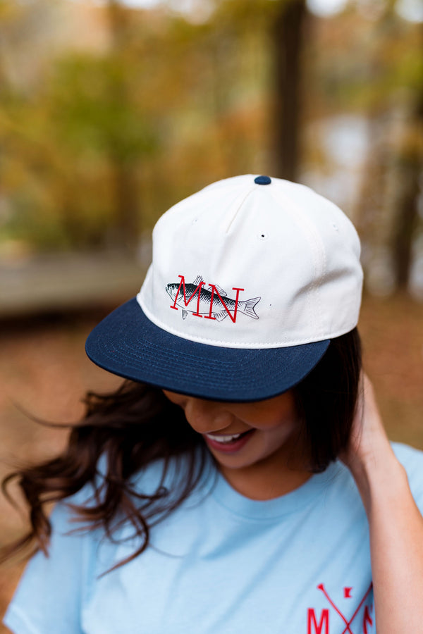 Person wearing a white and navy cap with 'MIN' and fish design, outdoors.