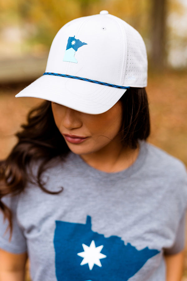 Woman wearing a white cap with a blue logo and a gray t-shirt with a blue design outdoors.