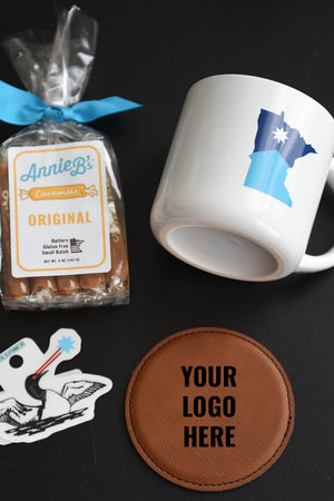 White mug with blue design, bag of caramels, coaster with 'Your Logo Here' text, and sticker on a black background