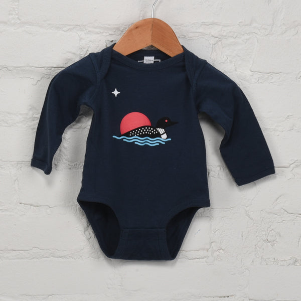 Navy blue baby onesie with a red sun and black loon design on a wooden hanger against a white brick wall.